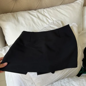 Svart kortkjol i enkel stil (shorts under) - En svart, stilren kortkjol, shorts under, med rak passform och diskreta sömmar framtill. Kjolen har en klassisk look och passar perfekt till sneakers eller boots. Materialet känns mjukt och följsamt, vilket gör den bekväm att bära hela dagen.