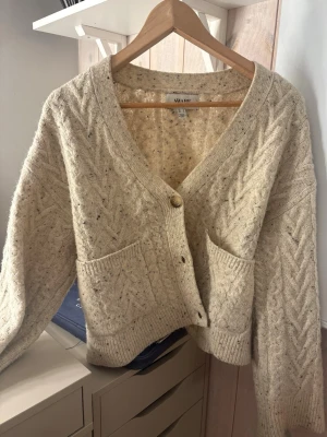 Beige stickad kofta från AWARE - Mysig beige stickad kofta från AWARE med grovt kabelstickat mönster och två stora fickor framtill. Koftan har v-ringning, ribbade muddar och knäpps med knappar. Perfekt för lager-på-lager och chill dagar.