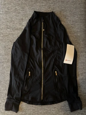 Svart zip-jacka från Lululemon - Snygg svart träningsjacka från Lululemon med guldfärgad dragkedja och två fickor med dragkedja. Figurnära passform och mjukt, stretchigt material som passar perfekt till träning eller som ett extra lager. Klassisk design med diskret logga på dragkedjan.