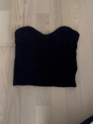 Svart ribbad stickad tubtopp - Svart stickad tubtopp med ribbad struktur. Toppen är axelbandslös och har en croppad passform, perfekt att styla med höga jeans eller kjol.
