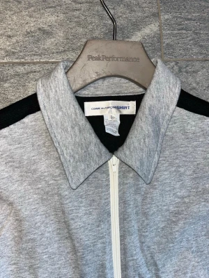 Grå zipjacka från Comme des Garçons - Grå jacka med svarta ärmar från Comme des Garçons SHIRT. Jackan har vit dragkedja framtill, klassisk krage och två fickor med dragkedja. Tillverkad i mjukt material, perfekt för en stilren och avslappnad look.