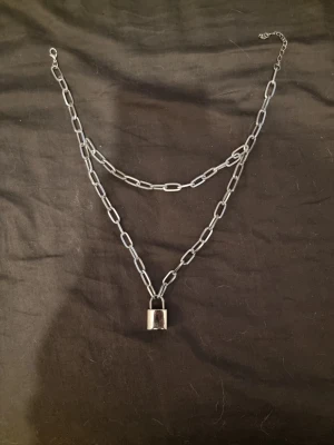 Silverfärgat halsband med hänglås - Coolt halsband i silverfärgad metall med två kedjor och ett hänglås som berlock. Kedjorna har stora länkar och ger ett edgy och trendigt intryck. Perfekt för att lyfta din outfit med en unik detalj.