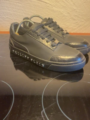 Svarta Philipp Plein sneakers - Snygga svarta sneakers från Philipp Plein med coola detaljer i ormmönstrad läderimitation. Skorna har en låg profil, svarta skosnören och märkets logga i silver på sidan av sulan.