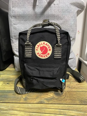 Svart Fjällräven Kånken ryggsäck - Klassisk svart Fjällräven Kånken ryggsäck Mini med ikonisk rund logotyp framtill. Väskan har randiga handtag och axelremmar i tyg samt en praktisk framficka med dragkedja. Tillverkad i slitstarkt material som tål vardagens äventyr. Höjd 27 cm Bredd 19 cm djup 9 cm