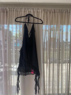 Svart omlottklänning med volang - Svart kortklänning med omlottmodell och volangdetaljer. Klänningen har tunna axelband som knyts i nacken och asymmetrisk nederkant med extra volanger. Perfekt för dig som gillar en lekfull och trendig stil.