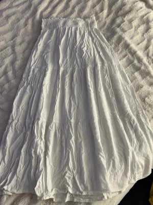 Vit långkjol med smock i midjan - Säljer en vit långkjol med smockad midja och flowig passform. Kjolen är i ett lätt och luftigt material, perfekt för varmare dagar. Enkel och stilren design som är lätt att matcha med olika toppar.