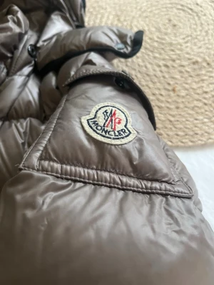 Brun dunjacka från Moncler med huva - Snygg brun dunjacka från Moncler med puffig design och huva. Jackan har dragkedja framtill, två sidofickor med dragkedja och klassisk Moncler-logga på ärmen. Perfekt för kalla vinterdagar och riktigt trendig look.