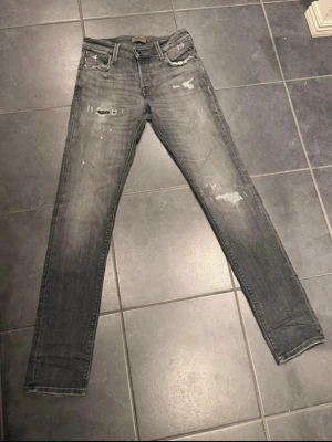 Grå skinny jeans Jack & Jones W27 L32 - Säljer ett par grå skinny jeans från Jack & Jones med slitna detaljer och distressed look. Jeansen har fem fickor, normal midja och är tillverkade i stretchig bomullsmix för skön passform. Perfekta för dig som gillar en trendig och avslappnad stil.