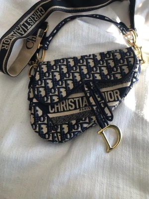 Saddle Bag blå - Ikonisk Saddle Bag i blått och beige canvas med monogrammönster. Väskan har guldfärgade metalldetaljer, bred axelrem och ett stort D-hänge framtill. Perfekt statement piece för dig som gillar lyxiga accessoarer.