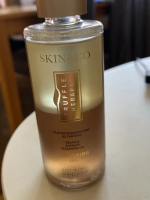 SKIN&CO Truffle Therapy Cleansing Oil - Lyxig ansiktsrengöringsolja från SKIN&CO i serien Truffle Therapy. Kommer i en genomskinlig plastflaska med guldfärgat lock och text i guld. Oljan är lätt gyllene och passar för normal till känslig hud. Tillverkad i Italien och innehåller tryffelextrakt.