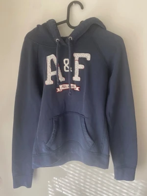 Mörkblå hoodie från Abercrombie & Fitch - Snygg mörkblå hoodie från Abercrombie & Fitch med stor broderad A&F-logga på bröstet och texten 'Ath Dept' under. Hoodien har huva med dragsko och en klassisk magficka. Den har krympt lite så det var en M men det är en S nu!