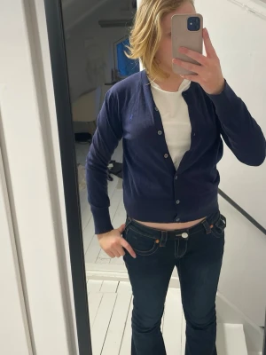 Mörkblå kofta från Polo Ralph Lauren - Säljer en mörkblå kofta från Polo Ralph Lauren med klassisk knappstängning och ribbade muddar. Koftan är tunn och har en diskret logga broderad på bröstet. Perfekt att slänga över en t-shirt för en chill look.