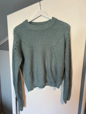 Grön stickad tröja med strukturmönster - Snygg stickad tröja i dämpad grön färg med strukturmönster och håldetaljer längs ärmar och framstycke. Tröjan har rund halsringning, långa ärmar och ribbade muddar. 