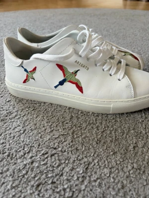 Vita Axel Arigato sneakers med broderi - Snygga vita sneakers från Axel Arigato med broderade fåglar i rött, grönt och blått på sidorna. Skorna är i lågt snitt och har vita skosnören samt en stilren design i läder. Perfekta för dig som gillar minimalistisk stil med en twist. Låda ingår ej. Nypris 2700!!