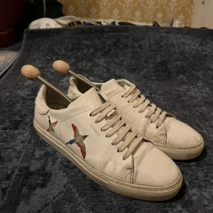 Axel Arigato skor - Vita sneakers från Axel Arigato i skinn med broderade fåglar på sidorna. Skorna har rund tå, snörning och en klassisk platt sula. Perfekta för dig som gillar stilrena detaljer och vill ha något unikt på fötterna. I storlek 44, hyfsat fint skick.