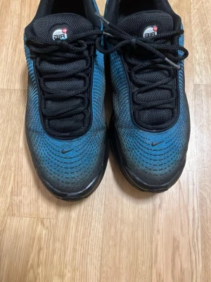 Nike Air Max Plus blå och svarta sneakers - Nike Air Max Plus sneakers i blått och svart med mesh-överdel och synliga Air-enheter i sulan. Skorna har svarta snören, diskret Nike-logga på tån och en sportig, futuristisk design. Perfekta för dig som gillar streetwear och vill sticka ut med en unik färgkombination.