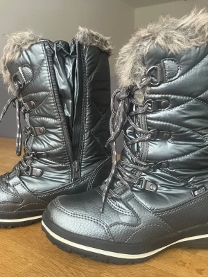 Svarta vinterstövlar med fuskpäls - Snygga svarta vinterstövlar från Everest med quiltad design, snörning och dragkedja på sidan. Stövlarna har grå fuskpäls upptill och är tillverkade i vattenavvisande material. Perfekta för kalla dagar och håller fötterna varma.