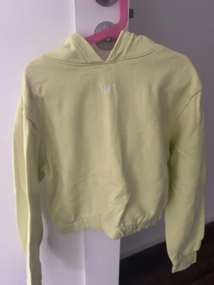 Gul hoodie med broderad NYC-text - Ljusgul hoodie med broderad vit NYC-text på bröstet. Modellen är croppad med resår i midjan och har en klassisk huva. Perfekt för en avslappnad och trendig stil. Mjuk bomullskänsla och långa ärmar.