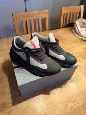 Svarta och grå Prada sneakers - Snygga sneakers från Prada i svart och grått med vita meshpaneler och svarta skosnören. Skorna har en tydlig Prada-logga på plösen och en chunky sula. Materialmix av läder och mesh ger en modern och sportig look.