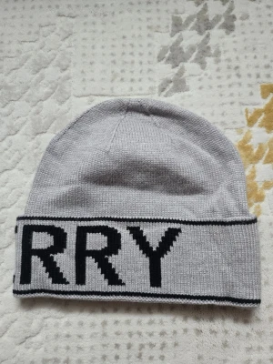Grå Burberry mössa med logga - Säljer en snygg grå mössa från Burberry med bred uppvikt kant och stor svart logotyp runtom. Mössan är stickad i mjukt material och har en klassisk, avslappnad passform. Perfekt för dig som vill ha en stilren accessoar med tydlig logga.