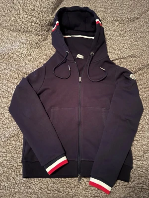 Mörkblå zip up hoodie från Moncler - Snygg mörkblå hoodie från Moncler med dragkedja, huva med dragsko och ribbade muddar. Detaljer i rött, vitt och blått på huva och ärmslut samt Moncler logga på ärmen. 