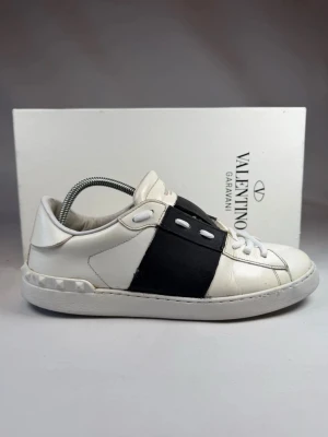 Valentino Garavani vita sneakers - Snygga vita sneakers från Valentino Garavani med bred svart läderdetalj över mitten och vita snören. Skorna har en låg profil och klassisk siluett med subtila nitar på hälen. Perfekta för dig som gillar stilrena och exklusiva sneakers.
