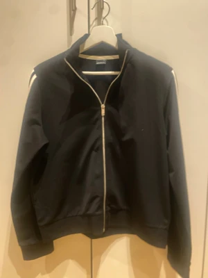 Svart track jacket med vita ränder - Svart track jacket med dragkedja framtill och ståkrage. Ärmarna har vita och mörkblå ränder längs sidorna. Jackan har ribbade muddar och är tillverkad i ett mjukt, sportigt material. Perfekt för träning eller chill.