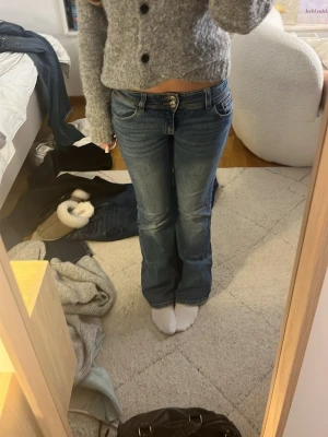 Blå bootcut jeans med låg midja - Säljer ett par klassiska blå bootcut jeans med låg midja och slitna detaljer. Jeansen har fem fickor, bälteshällor och stängs med knapp och dragkedja. Perfekta för en avslappnad och trendig look.