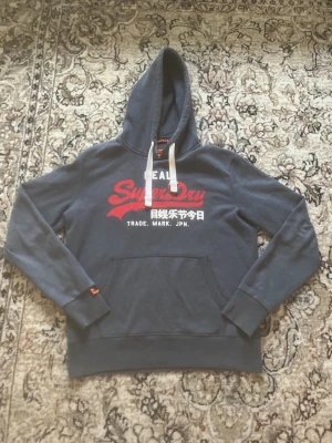 Superdry hoodie - Säljer min superdry hoodie använd ett fåtal gånger. Ny pris är 799kr men den finns inte längre (vintage). Endast några fåtal defekter 3st som visas på de sista bilderna lite slitet vid kragen och en del av texten och en annan del av texten som har en svart liten prick (fabriksdefekt). Annars top skick 9/10. Skriv till mig om ni vill ha mått eller undrar något!