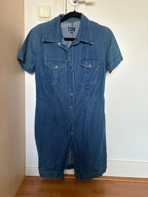 Blå jeansklänning från VNL - Säljer en blå jeansklänning från VNL med korta ärmar, knappar framtill och två bröstfickor.  Väldigt fin till sommaren!