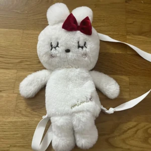 Vit plyschkanin-väska med rosett - Supersöt väska i form av en vit kanin med mjuk plysch, broderade ögon och en röd sammetsrosett på huvudet. Väskan har vita axelband och små pärldetaljer vid halsen. Perfekt accessoar för dig som gillar gulliga detaljer och unik stil.