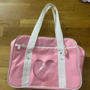 Rosa handväska med hjärtfönster - Söt rosa handväska med vita detaljer och axelband. Väskan har ett genomskinligt hjärtfönster på framsidan och en liten dragkedjeficka. Dekorerad med pärlor på ena handtaget. Perfekt för dig som gillar gullig och lekfull stil.