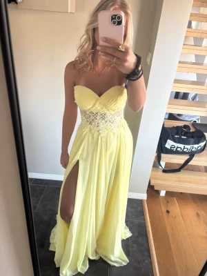 Ljusgul axelbandslös långklänning - Säljer en ljusgul axelbandslös långklänning med v-ringad överdel och spetsdetaljer i midjan. Klänningen har en hög slits på ena sidan och ett svepande, flowy tyg som ger en elegant look. Perfekt för bal eller student.