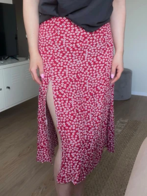 Blommig långkjol med slits och dragkedja- stl M, mycket bra skick - Säljer en röd långkjol med vita blommor och en slits vid vänster ben. Lätt, luftig och perfekt flowy. Så fin till en liten topp och kanske en jeansjacka nu till vår och sommar!