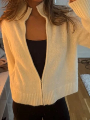 Vit stickad ziptröja - Mysig vit stickad ziptröja med hög krage och långa ärmar. Tröjan har en rak passform och ribbstickade muddar vid ärmslut och nederkant. Perfekt att dra på sig när det är kyligt ute.