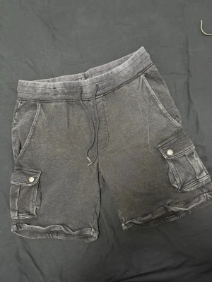 Svarta cargoshorts med snörning - Säljer ett par svarta cargoshorts med snörning i midjan och flera fickor med knappstängning på sidorna och bak. Shortsen har en avslappnad passform och är perfekta för chill dagar. Materialet känns mjukt och bekvämt, troligen bomull eller bomullsblandning.