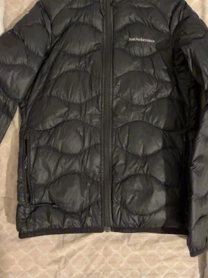 Svart dunjacka Peak Performance - Snygg svart dunjacka från Peak Performance med quiltat mönster och dragkedja framtill. Jackan har två sidofickor med dragkedja och diskret logga på bröstet. Perfekt för kalla dagar och enkel att matcha med olika outfits.