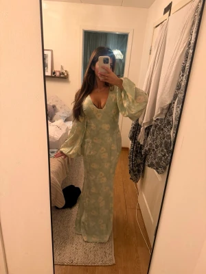 Ljusgrön blommig långklänning från adoore - Säljer en ljusgrön långklänning med blommigt mönster och v-ringad front från adoore. Klänningen har vida, långa ärmar och en elegant passform som framhäver figuren. Perfekt för sommarens alla tillfällen! Prislapp sitter kvar! I storlek L men är liten i storlek, passar mig som har S.