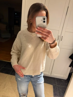 Vit stickad tröja med pärlor - Säljer en vit stickad tröja med pärldetaljer över hela framsidan och ärmarna. Tröjan har rund halsringning, ribbade muddar och en något boxig passform. Perfekt för dig som gillar en söt och trendig look.