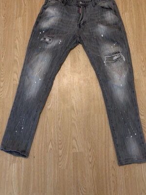 Mörkgrå dsq 2 jeans - Säljer ett par grå jeans med coola slitningar och färgstänk på både fram- och baksida. Jeansen har raka ben, klassisk femficksmodell och detaljerade revor på låren. Perfekta för dig som gillar en avslappnad och trendig streetstil.