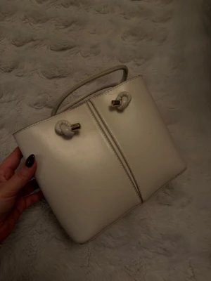 Krämvit handväska med gulddetaljer - Snygg krämvit handväska i slätt konstläder med guldfärgade detaljer och dekorativa knutar på handtagen. Väskan har både handtag och axelrem samt ett praktiskt innerfack med dragkedja. Perfekt för dig som gillar stilrena accessoarer.