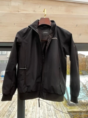 Svart vindjacka Peak Performance - Snygg svart vindjacka från Peak Performance med hög krage och dragkedja framtill. Jackan har två framfickor med dragkedja och ribbade muddar vid ärmslut och nederkant. Tillverkad i ett lätt och slitstarkt material, perfekt för blåsiga dagar. Storlek 160. Mycket bra skick