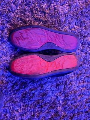 Svarta sneakers från Christian Louboutin - Säljer ett par svarta sneakers från Christian Louboutin med klassisk röd sula och signaturtext undertill. Skorna har svart mocka, svarta skosnören och vit insida. Perfekta för dig som vill ha något stilrent men ändå lyxigt. Loubs 