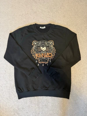Svart sweatshirt med tiger från Kenzo - Svart sweatshirt från Kenzo med stor broderad tiger och texten 'KENZO PARIS' på bröstet. Tröjan har rund hals, ribbade muddar och är långärmad. Perfekt för dig som gillar streetwear och vill ha en ikonisk detalj i garderoben. Fråga gärna om du har några frågor!