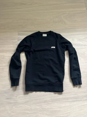 Svart sweatshirt från Nudie Jeans - En stilren svart sweatshirt från Nudie Jeans i 100% ekologisk bomull. Tröjan har rund halsringning, långa ärmar och en liten vit logotyp på bröstet. Perfekt för en avslappnad och trendig look.