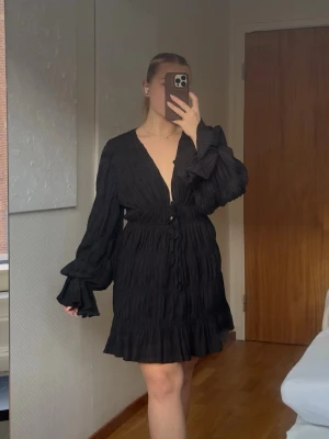 Svart kortklänning NA-KD x Moa Mattsson - Svart kortklänning från Moa Mattssons kollektion med NA-KD. Den har v-ringning, snörning i midjan som går att justera och volangdetaljer vid ärmslut och nederkant. Klänningen har långärmade ärmar och är tillverkad i ett lätt, krusat material. 