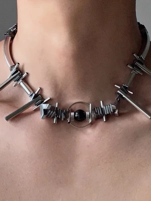 Silvrigt titanium steel statement halsband med pärla - Unikt halsband i silverfärgad metall med geometriska former och en svart pärla i mitten. Designen är edgy och modern med flera stavar och trianglar som ger en cool look. Perfekt för dig som vill sticka ut och addera något extra till din outfit. brand: Illuminory