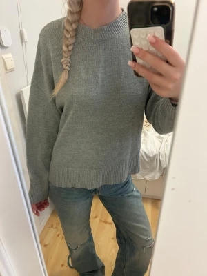 Grå stickad tröja med rund hals - En enkel och stilren grå stickad tröja med rund halsringning och långa ärmar. Tröjan har ribbade muddar vid ärmslut och nederkant, vilket ger en avslappnad men snygg look. 