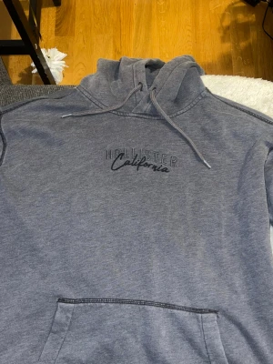 Grå hoodie från Hollister - Snygg grå hoodie från Hollister med broderad text 'Hollister California' på bröstet. Hoodien har en klassisk känguruficka framtill och justerbar huva med dragsko. Tillverkad i mjukt bomullsmaterial som är skönt mot huden. Perfekt för en avslappnad och trendig look.