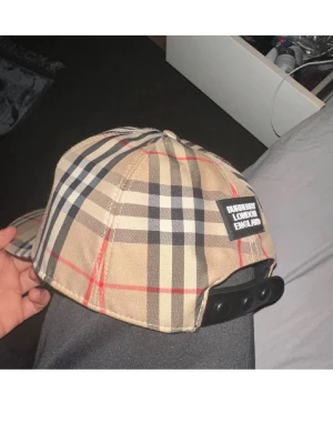 Burberry beige rutig keps - Säljer en klassisk Burberry keps i beige med det ikoniska rutmönstret i svart, vitt och rött. Kepsen har justerbar snapback och en svart patch med texten 'Burberry London England' bak. Perfekt accessoar för en trendig och stilren look.